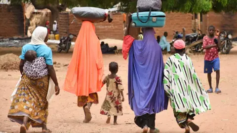 AFP via Getty Images Three women and several children, with their backs to the camera, leave Kukawa village in April 2022 after their houses were burnt down. Two of the women, wearing colourful robes, have luggage on their heads.