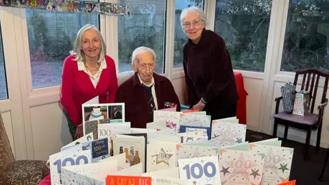 Tony Winterburn is sitting at a table full of birthday cards. Two women are standing next to him, smiling at the camera.