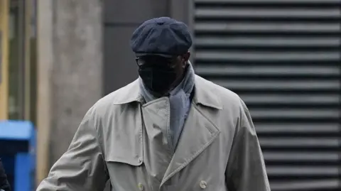 Charles Ehikioya is wearing a beige trench coat, grey flat cap, black face mask, glasses and a light grey scarf is seen walking outdoors.