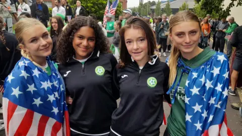 Four young female soccer players stand side by side. They are wearing team colours and are draped in the US flag.