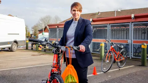 Beccy Marston pictured holding a red bike. She has a brown pixie haircut, wears a grey shirt underneath a blue blazer.