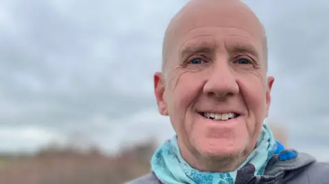 Emma Baugh/BBC Richard Astle standing outside. He had an aqua coloured scarf around his neck and is wearing a dark quilted zipped coat. He is smiling broadly. Behind him are fields, shrubs and grey sky. 