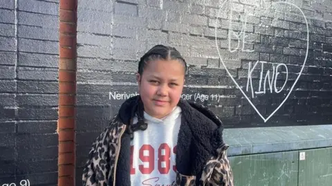 A young girl with dark hair tied in braids looks towards the camera. She is wearing a hooded leopard print jacket with a black fluffy lining, on top of a white top. She standing in front of a black painted brick wall that has the words be kind written on it inside a heart