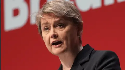 PA Media Headshot of Home Secretary Yvette Cooper speaking at the Labour Party's autumn conference in Liverpool last month. She is wearing a dark jacket and her grey hair is styled in a pixie cut. She has small earrings and stands against a bright red backdrop.