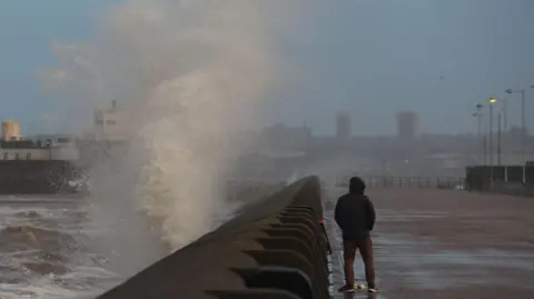 EPA Waves crash against the sea wall in the UK as a man stands nearby