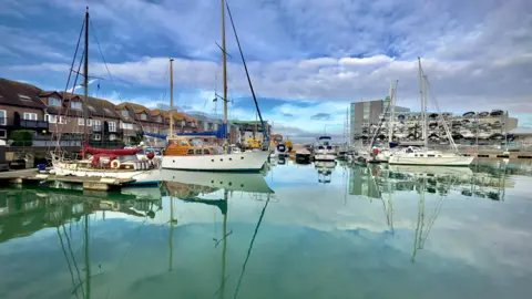 Several boats are moored up in the harbour. The water is a beautiful green colour and is so crystal clear that it offers a perfect reflection of the boats and blue sky above them. 