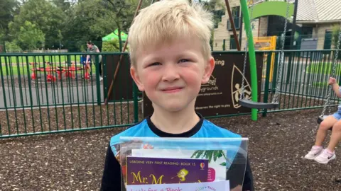BBC Theo, een jongen met blond haar en een blauw shirt houdt zijn boeken omhoog