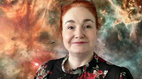 A woman with red hair and glasses on her head stands in front of a galaxy-inspired backdrop. She is wearing a black dress with a floral design on.