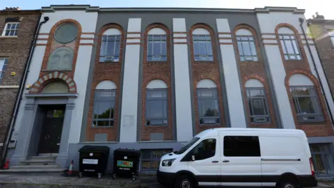 An imposing terrace building of red brick with grey and white painted detail. There are two rows of arched windows to the right of a tall arched door. There are large rectangular black bins and a white van out front. In the top left corner of the facade is a circular window with the Star of David etched into it.