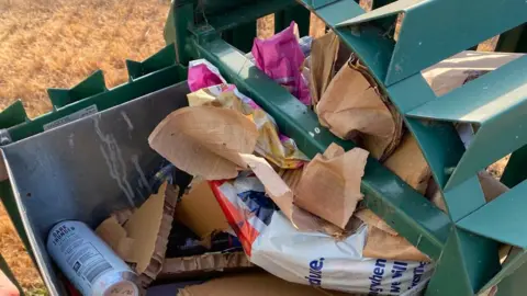 Cullompton Town Council An overflowing green coloured council bin in a park. There is lots of items of cardboard and plastic bags in there. 