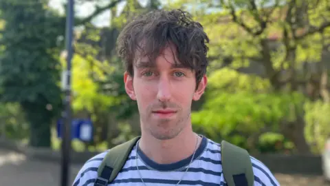 BBC/Elizabeth Baines A man with brown hair and blue eyes wears a navy and white striped top. He has green backpack straps on his shoulders and a silver necklace. Behind him, trees are blurred.