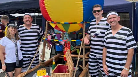 Paul Beesley, Lee Masser and Jon Chaproniere wearing black and white-striped prisoner outfits, with Jo Chaproniere dressed as a police officer. They are standing next to a large brown kart with a colourful hot air balloon on top.