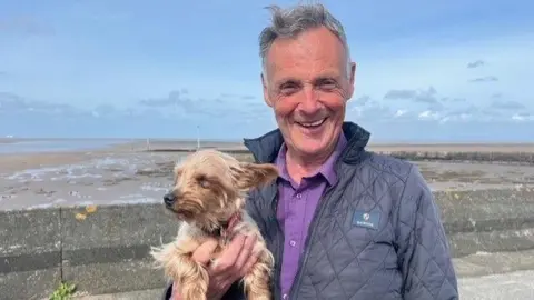 Gayle Harmsworth wears a purple polo shirt and quilted navy-blue jacket. He has a small brown terrier in his arms - Lola.