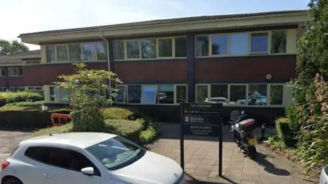 A two-storey red-brick office block, with yellow window frames and a sign outside reading "The Parkway" and "Hampshire County Council". A white car is parked on the road outside