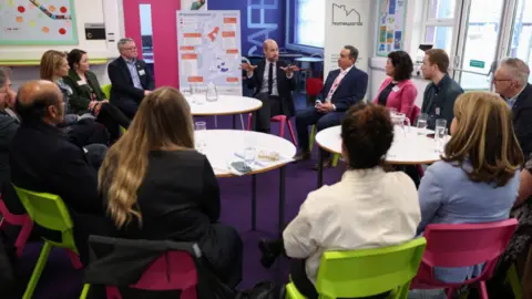 PA Media The Prince of Wales surrounded by people in smart clothing sitting on pink and green chairs in a school