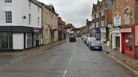 Google A high street with shops on either side of the road and a row of parked cars to the right.