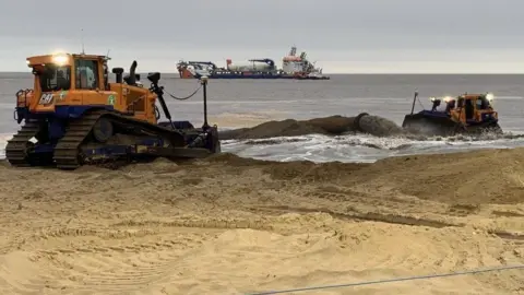 Environment Agency Heavy plant machinery replenishing sand on a beach. There is a large ship in the sea in the background.