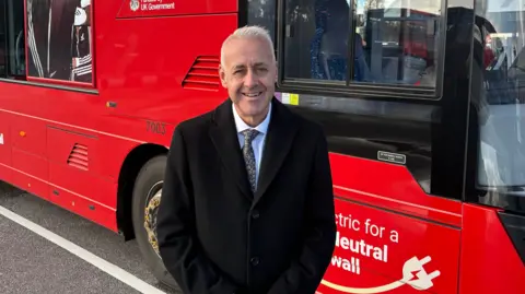 Richard Stevens wearing a black coat with a white shirt and multi coloured tie standing in front of a red double decker bus