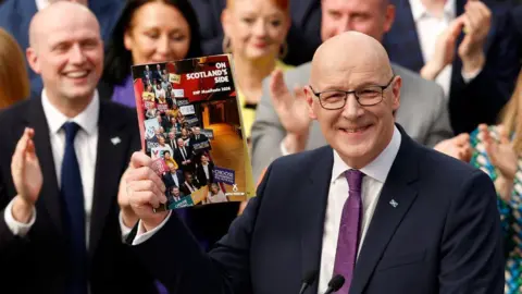 John Swinney, who is bald with glasses, holds up a manifesto booklet while people applaud behind him. He is smiling at the camera 