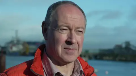A man with short grey hair standing next to a body of water with out of focus buildings behind. He is wearing a red coat and a pink shirt. It is a sunny day.