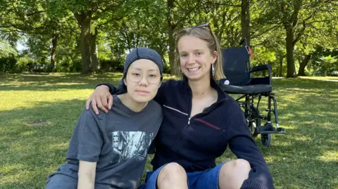 Sofía Luis-Hobbs/BBC Two women sat in the shade in a park on a bright day. The girl on the right has shoulder length blonde hair and wears a black zip up jumper and denim shorts as she puts her arm around her friend Crystal who sits to her left. Crystal wears a grey graphic t-shirt, glasses, and wears a bandana in her hair. There is an empty wheelchair in the background.