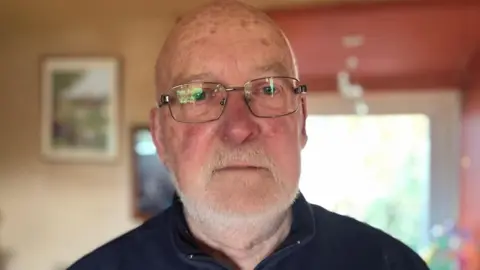 An older man staring, not smiling at the camera. He's wearing a dark shirt, has a grey beard, bald head, freckles and glasses