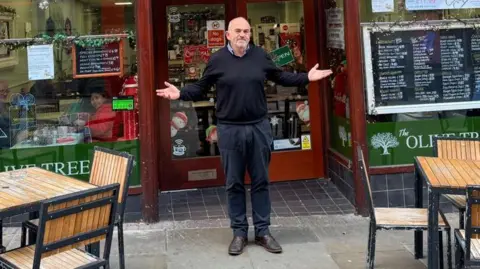 Kevin in front of his cafe. It has green signage and there are tables and chairs at the side of him. He has his hands up towards the sky. 