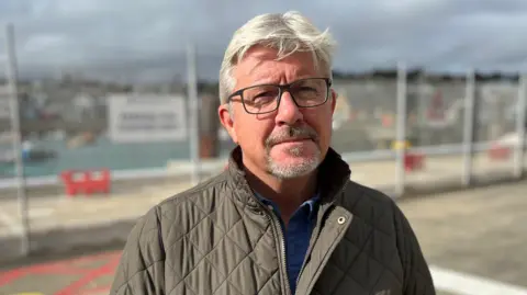 A man with grey hair, black glasses and a grey beard and sole patch. He's wearing a green Barbour jacket.