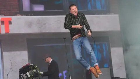 A man is holding a microphone while jumping in to the air. He is wearing blue jeans and a striped shirt.