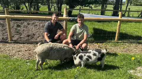 Two men with dark curly hair and dark facial hair are crouching on some grass with farm enclosures behind them. On the grass with them are two small pigs. One is a grey and beige colour and the other is black and white