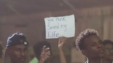 Aswat Almadina Man holding a sheet of paper with a handwritten note that reads your music saved my life. on the right of the picture are two other men. They are part of a crowd and only their heads are pictured.