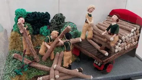 Several knitted characters in brown overalls lift logs into the back of a lorry.