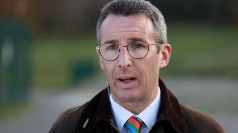 Andrew Muir has short greying hair and is wearing glasses. He is wearing a brown jacket over a white shit with a multi-coloured tie.