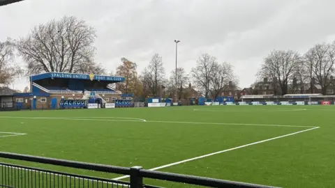 Owen Hardwick, BBC An artificial pitch dominates the foreground, with a small football stand in the left side of the background with the words "Spalding United Football Club" printed on top. It is a cloudy day with trees bare.