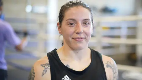 Jamie Niblock/BBC Eilish Tierney smiles at the camera. She wears a black adidas vest and has tattoos on both arms and shoulders - the visible ones are of Cupid flying with an arrow. Her brown hair is tied back and she has a silver nose ring. 