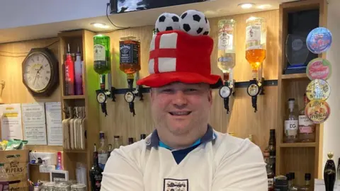 Sean Rowbottom is standing behind the bar and smiling brightly at the camera. He is wearing a tall red hat with an England flag on it and three fluffy football balls on top. He has stubble and is wearing a white England kit.