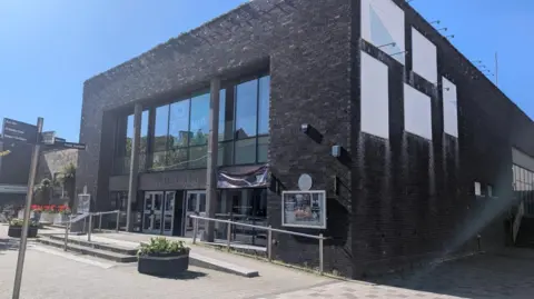 A general view of the entrance to the Civic Hall in Whitehaven. It is a one-storey building with a flat roof, built in dark bricks, with a glass-fronted entrance.