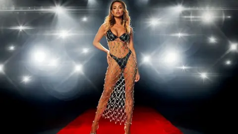 ITV Georgia Harrison, with her left hand on her hip, she is wearing a black bikini, with a mesh chainmail dress over the top.