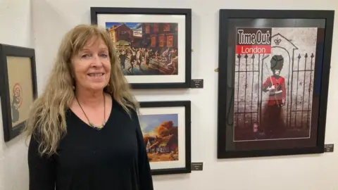 James Grant/BBC Woman with long light brow hair wearing black stands in front of pictures on a wall. One shows a guardsman.