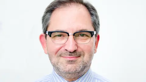 A man with dark hair and a greyish beard with dark framed glasses and a blue and white striped shirt. He looks into the camera with a white wall behind him.