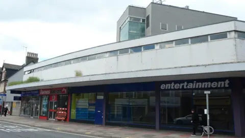 Christine Johnstone/Geograph Crossways shopping centre in Paignton. It consists of large grey buildings with empty shop units on the ground floor. People are walking along the street.