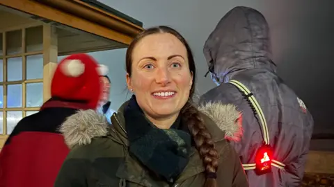 Katie smiles at the camera as three men stand behind her. She has a green coat on with a fake fur lining around the hood. She has a dark blue scarf as well. Her long dark hair is platted into a ponytail which is over her right shoulder.