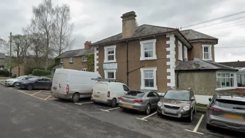 A two storey building with a brown cobbled exterior and white window pains. There are cars parked in the car park alongside it. 