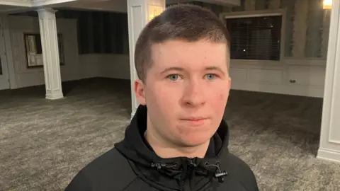 Conor looking at the camera. He has short dark hair and is wearing a black jacket.