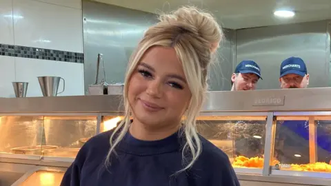 Blonde woman looking at the camera from behind a chip shop counter with display cabinets visible containing fish and other battered goods 