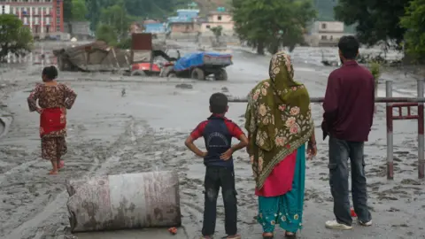 The backs of two women, a man and a boy looking out to partially submerged trucks and buildings as mud covers a section of a highway in Nuwakot in central Nepal