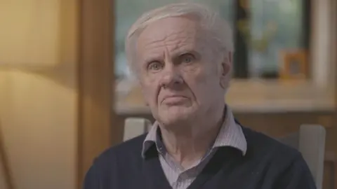 A man with grey, thinning hair looks at the camera. His hair is swept to the side as he sits on a wooden backed chair. The man is wearing a dark jumper and chequered shirt, with an open collar. Behind him is a blurred view of a window or bright painting, surrounded by yellow walls. 