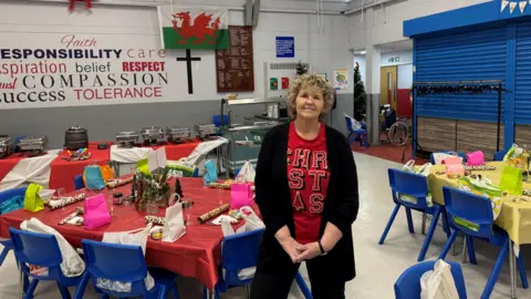 Carolyn wearing a red Christmas top with a black cardigan, smiling with short blonde curly hair, standing with tables set ready for Christmas dinner with red table cloths and Christmas crackers