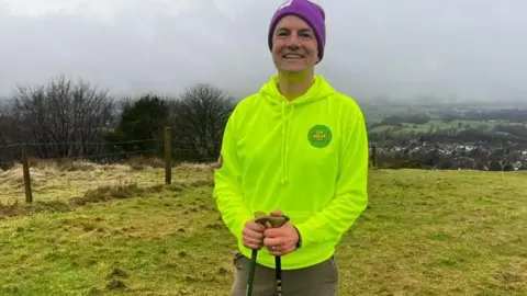 A man wearing a neon green hoodie and purple beanie hat, and brown trousers, smiles at the camera as he holds two black walking sticks, and stands in a green field, with rolling fields as a view behind him. It is a cloudy, foggy day