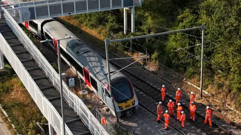 PA Media A photograph of the Greater Anglia train with part of the train off the track at Wickford in Essex. The train has been stopped and is not moving. Around 10 or more people are wearing orange fluorescent hi-vis with white helmets and are stood on the tracks to the right of the train.
